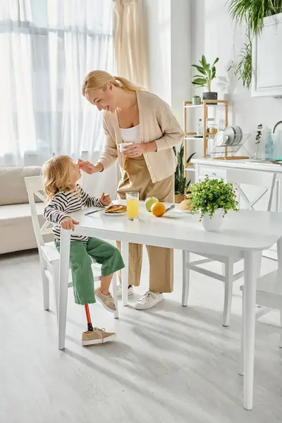 Happy mother standing near daughter with prosthetic leg having breakfast in kitchen, adaptive — Stock Photo