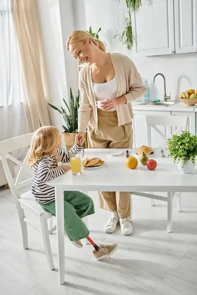Happy mother standing near cute daughter with prosthetic leg having breakfast in kitchen, inclusion — Stock Photo
