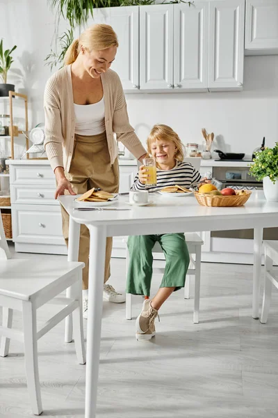 Happy woman serving pancakes to daughter with prosthetic leg during breakfast in kitchen, inclusion — Stock Photo
