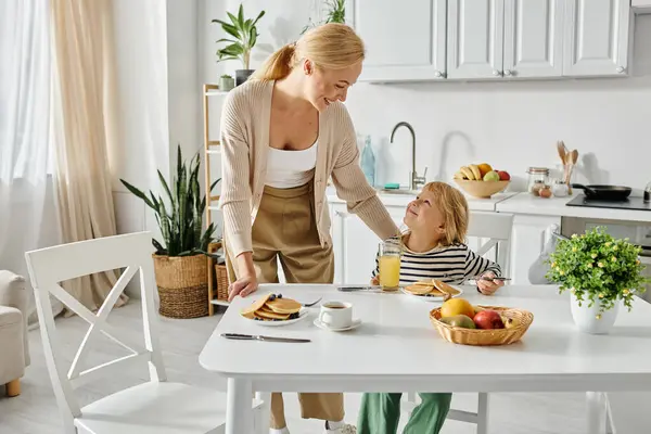 Happy mother looking at cute daughter with prosthetic leg during breakfast in kitchen, inclusion — Stock Photo