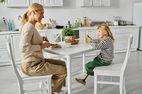 Mãe feliz tomando café da manhã com sua filhinha com perna protética na cozinha, inclusão — Fotografia de Stock