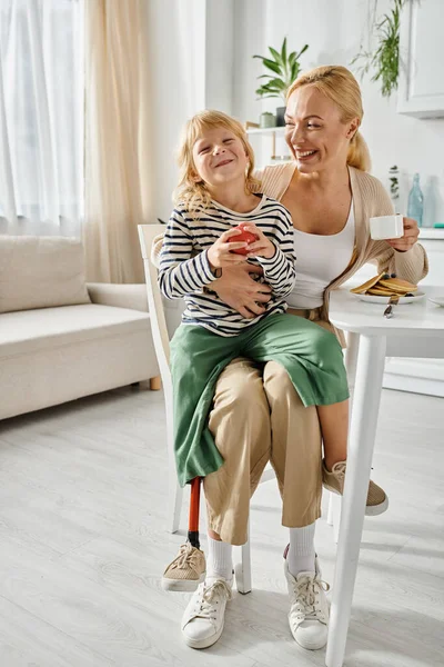Preschooler girl with prosthesis leg sitting on laps of happy mother during breakfast in kitchen — Stock Photo