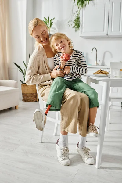 Bambina con protesi seduta sulle ginocchia di madre felice durante la colazione in cucina — Foto stock