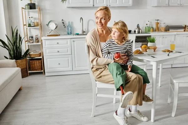 Ragazza carina con gamba protesica seduta sulle ginocchia di madre felice durante la colazione in cucina — Foto stock