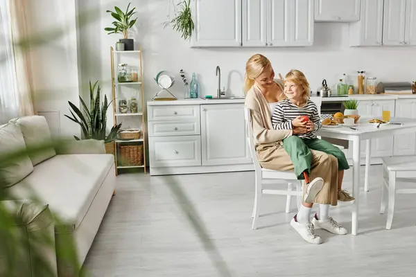 Little girl with prosthetic leg sitting on laps of happy blonde mother during breakfast in kitchen — Stock Photo