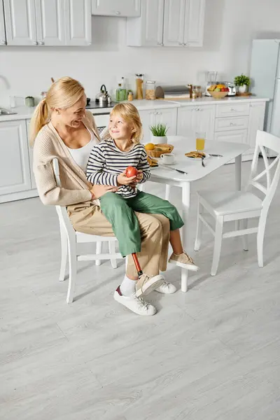 Preschooler girl with prosthetic leg sitting on laps of happy mother during breakfast in kitchen — Stock Photo