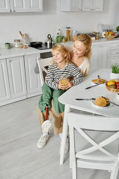 Bambino in età prescolare con protesi sulla gamba seduta sulle ginocchia di madre felice durante la colazione in cucina — Foto stock