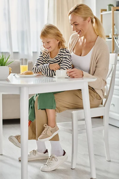 Bambino con gamba protesica seduto sulle ginocchia di madre felice e guardando frittelle durante la colazione — Foto stock