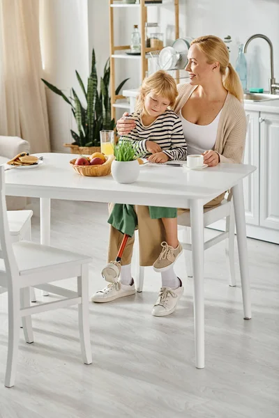 Girl with prosthetic leg sitting on laps of happy mother and looking at pancakes during breakfast — Stock Photo