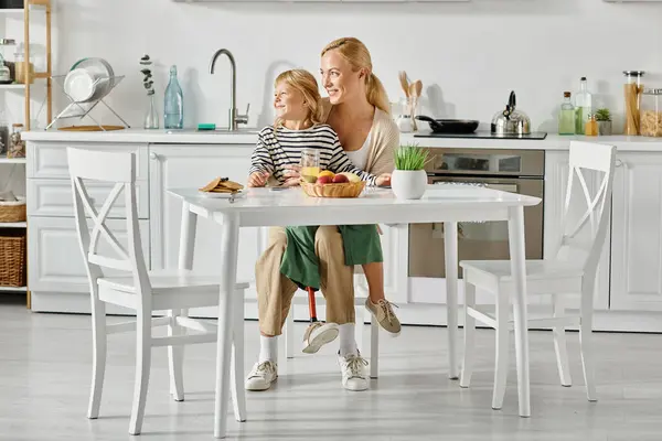 Preschooler girl with prosthetic leg sitting on laps of happy mother during breakfast in kitchen — Stock Photo