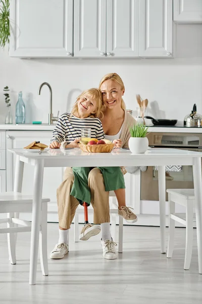 Cute kid with prosthetic leg sitting on laps of happy mother during breakfast in kitchen — Stock Photo