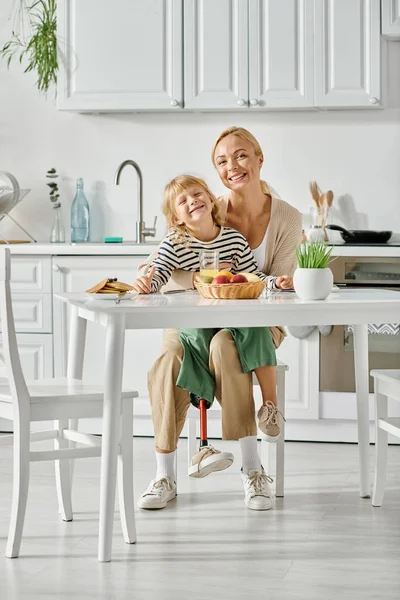 Cheerful girl with prosthetic leg sitting on laps of happy mother during breakfast in kitchen — Stock Photo