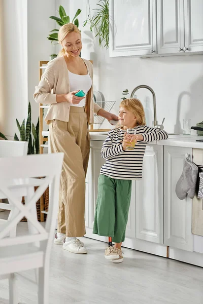 Mädchen mit Beinprothese hält ein Glas Orangensaft in der Nähe der glücklichen Mutter beim Geschirrspülen in der Küche — Stockfoto