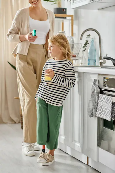 Fille avec prothèse jambe tenant verre de jus d'orange près de mère heureuse laver la vaisselle dans la cuisine — Photo de stock