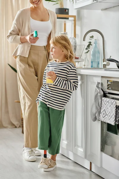 Girl with prosthetic leg holding glass of orange juice near happy mother washing dishes in kitchen — Stock Photo