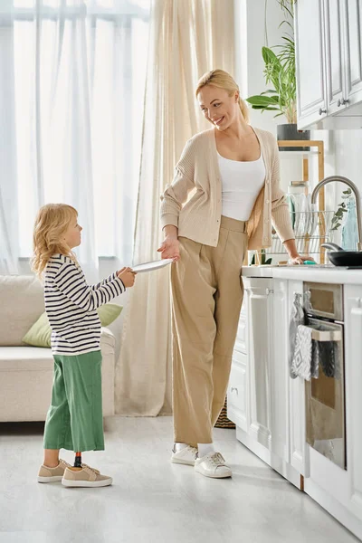 Nettes Mädchen mit Beinprothese hält Teller und hilft glücklicher Mutter beim Geschirrspülen in der Küche — Stockfoto