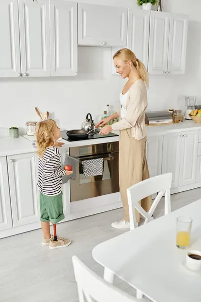 Мила дівчина з протезною ногою тримає тарілку і допомагає щасливій мамі мити посуд на кухні — стокове фото