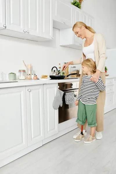 Радостная мама готовит и держит за руку маленькую дочь-инвалида с протезной ногой на кухне — стоковое фото