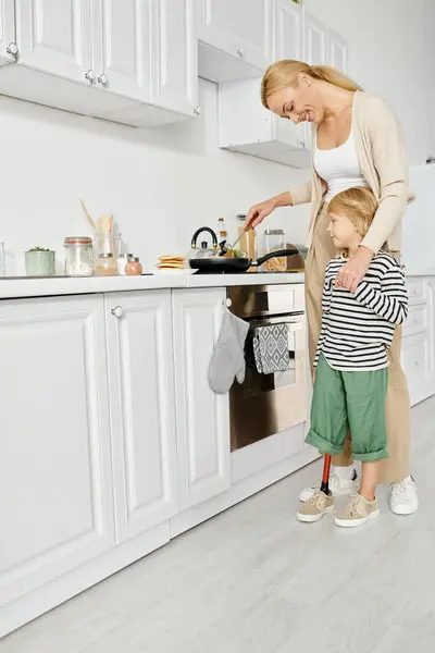 Счастливая мама готовит и держит за руку маленькую дочь-инвалида с протезом ноги на кухне — стоковое фото