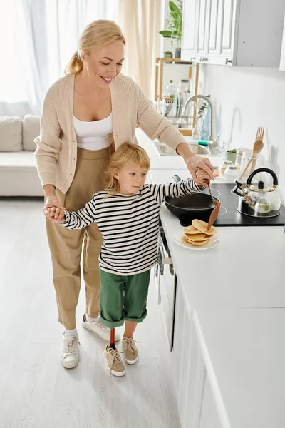 Feliz madre de pie cerca de linda hijita con prótesis pierna fritura panqueques en la cocina - foto de stock