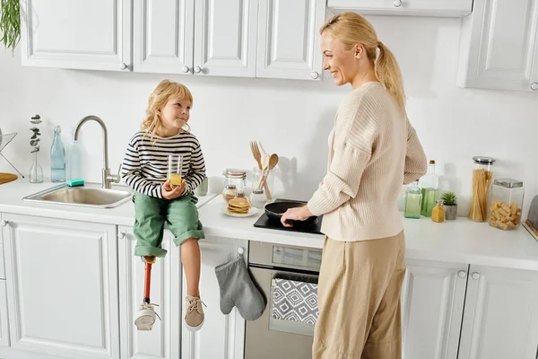 Happy girl with prosthetic leg sitting on countertop with orange juice near mother cooking pancakes — Stock Photo