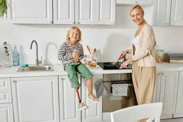 Fille heureuse avec la jambe prothétique assis sur le comptoir avec du jus d'orange près de la mère crêpes de cuisson — Photo de stock
