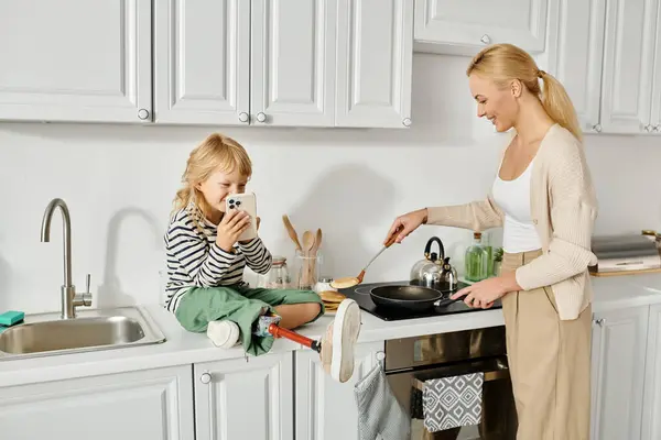 Happy girl with prosthetic leg sitting on countertop with smartphone near mother cooking pancakes — Stock Photo