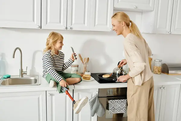 Niña con la pierna protésica sentada en la encimera y mostrando el teléfono inteligente a la madre cocinar panqueques - foto de stock
