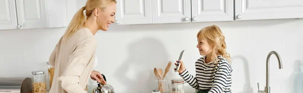 Glückliches kleines Mädchen, das auf der Arbeitsplatte sitzt und Mutter das Smartphone beim Kochen in der Küche zeigt, Banner — Stockfoto