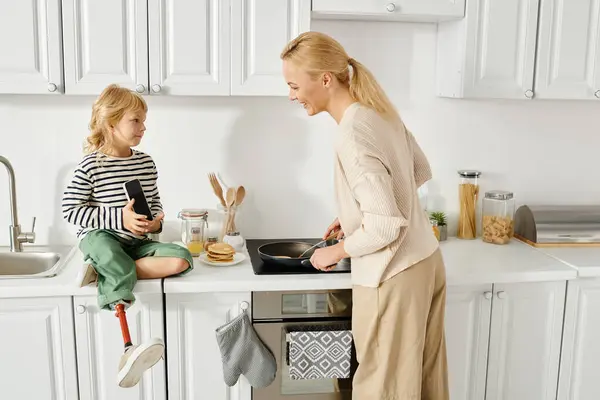 Happy girl with prosthetic leg sitting on countertop with smartphone near mother cooking pancakes — Stock Photo