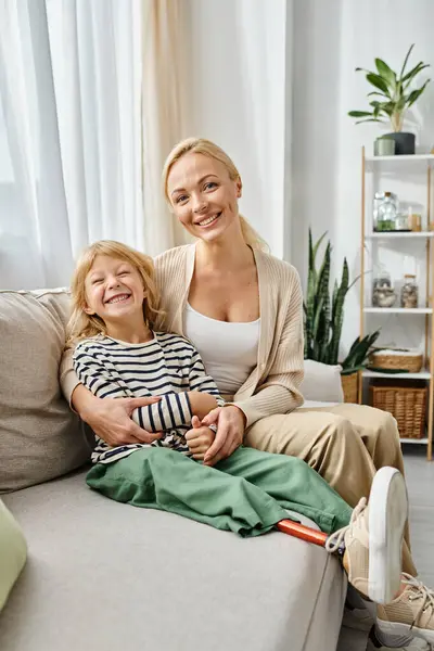 Mãe feliz abraçando filha com prótese perna e sentados juntos no sofá na sala de estar — Fotografia de Stock