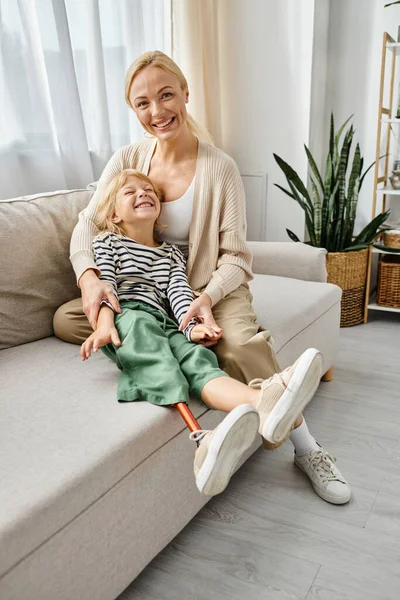 Happy mother hugging cute daughter with prosthetic leg and sitting together on sofa in living room — Stock Photo