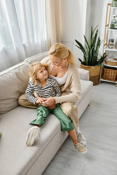 Madre rubia abrazando al niño feliz con la pierna protésica y sentados juntos en el sofá en la sala de estar - foto de stock