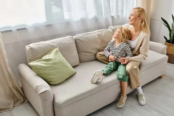 Feliz madre abrazando a la hija con la pierna protésica y sentados juntos en el sofá en la sala de estar - foto de stock
