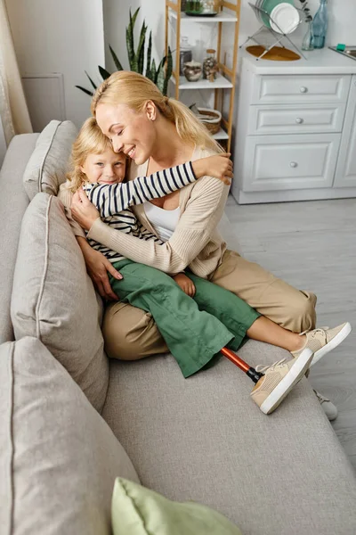 Mutter umarmt glückliche Tochter mit Beinprothese und sitzt zusammen auf Sofa im Wohnzimmer — Stockfoto