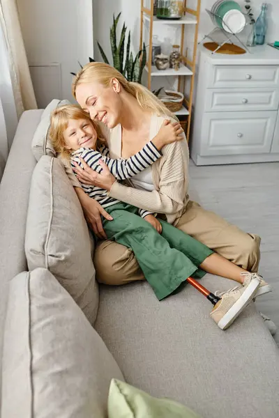 Allegra madre che abbraccia la figlia con una gamba protesica e si siede insieme sul divano in soggiorno — Foto stock
