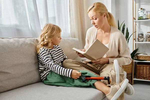Mujer rubia leyendo libro a hija con prótesis de pierna mientras están sentados juntos en la sala de estar - foto de stock