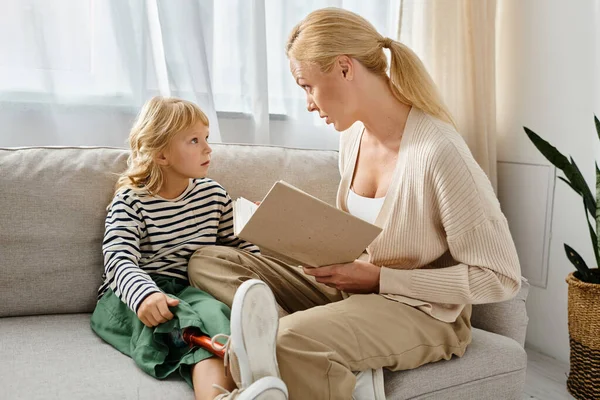 Blonde mother reading book to little girl with prosthetic leg while sitting together in living room — Stock Photo