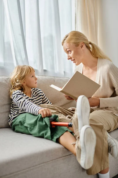 Rubia madre lectura libro a linda hija con prótesis pierna y sentado juntos en sala de estar - foto de stock