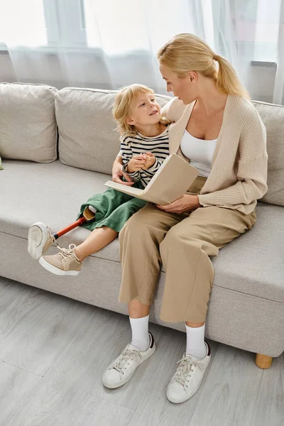 Rubia madre lectura libro a feliz hija con prótesis pierna y sentado juntos en sala de estar - foto de stock