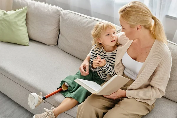 Blonde Mutter liest ihrem Kind mit Beinprothese Buch vor, während sie zusammen im Wohnzimmer sitzt — Stockfoto
