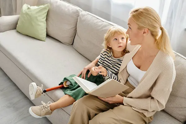 Rubia madre lectura libro a lindo niño con prótesis pierna mientras están sentados juntos en sala de estar - foto de stock