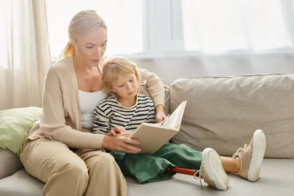 Madre rubia leyendo libro a su hijo con la pierna protésica mientras están sentados juntos en la sala de estar - foto de stock