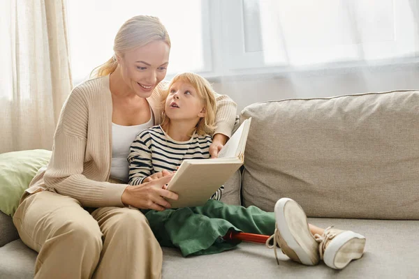 Feliz madre leyendo el libro a su hija con la pierna protésica y sentados juntos en la sala de estar - foto de stock