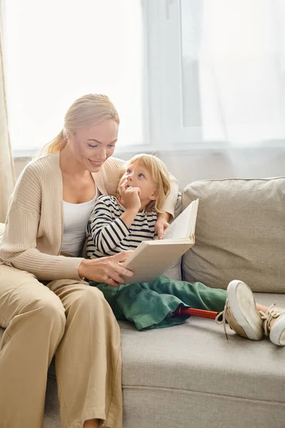 Feliz madre leyendo el libro a su hijo rubio con la pierna protésica, sentados juntos en la sala de estar - foto de stock