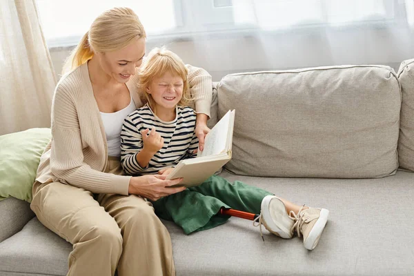 Mère lisant le livre à sa fille heureuse avec la jambe prothétique et assise ensemble dans le salon — Photo de stock