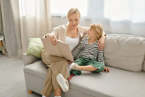 Madre leyendo libro a su hija alegre con la pierna protésica y sentados juntos en la sala de estar - foto de stock