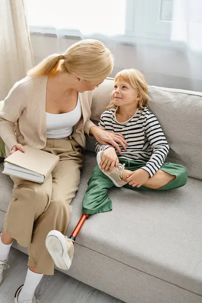 Glückliches Mädchen mit Beinprothese sitzt auf Sofa neben blonder Mutter mit Buch im Wohnzimmer — Stockfoto