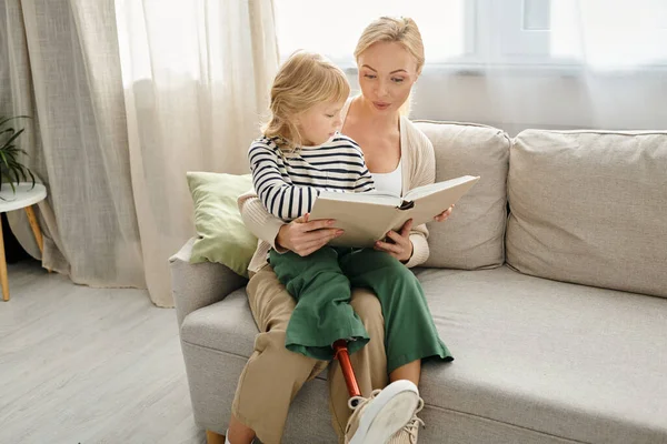 Glückliches Mädchen mit Beinprothese sitzt auf dem Schoß einer blonden Mutter und liest Buch im Wohnzimmer — Stockfoto