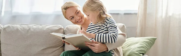Cheerful girl sitting on laps of blonde mother and reading book together in living room, banner — Stock Photo